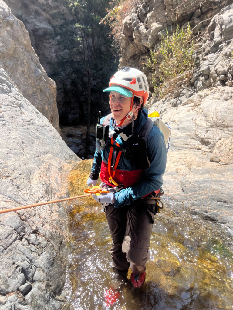A person dressed in a white helmet with orange accents, teal cap, blue jacket, and gloves is smiling while holding an orange rope and rappelling down a rocky canyon with shallow water at their feet. The background shows steep rock walls and some vegetation in Eaton Canyon.