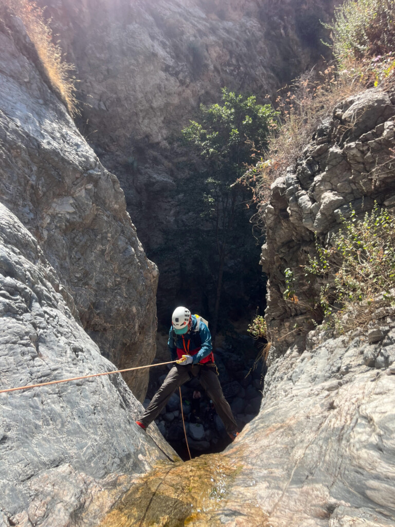A person equipped with a white helmet, gloves, and outdoor clothing is rappelling down a steep rocky canyon wall using a rope. The surrounding area features rugged rock formations and sparse vegetation under bright sunlight. The scene captures the adventurous activity of canyoneering in Eaton Canyon.