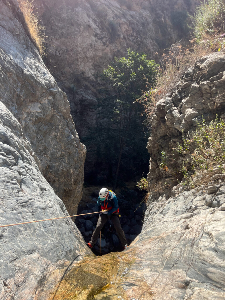 A person equipped with a white helmet, gloves, and harness is rappelling down a steep rocky canyon wall using an orange rope. The surrounding area features rugged rock formations, sparse vegetation, and a small trickle of water flowing down the rock surface. Sunlight illuminates parts of the canyon, highlighting the texture of the rocks and greenery.