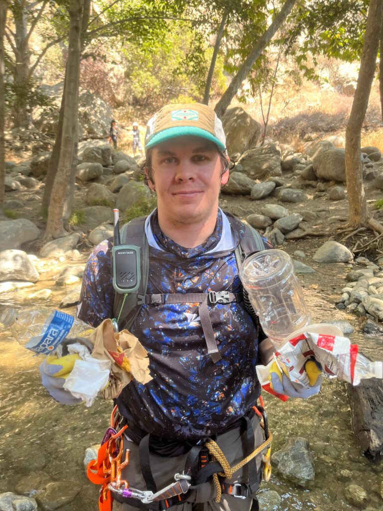 A man dressed in canyoneering gear and a cap stands in a rocky creek surrounded by trees, holding various pieces of trash such as plastic bottles and wrappers in both hands, participating in a cleanup activity during a canyoneering trip in Eaton Canyon.