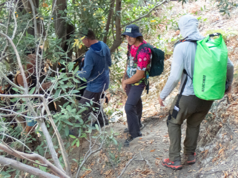 Several people hiking through a dense forest trail in Eaton Canyon. One person in the foreground wears a light gray hoodie and carries a bright green waterproof backpack. Another person wears a colorful shirt with a dark bucket hat and a dark green backpack. The group is surrounded by green foliage and dry leaves on the ground, indicating a natural, rugged outdoor environment.