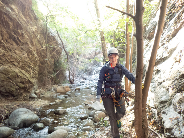 A person dressed in canyoneering gear including a helmet, harness, gloves, and ropes stands next to a tree in a rocky canyon with a shallow stream and scattered rocks. The canyon walls are steep and lined with trees and greenery, illuminated by bright sunlight filtering through the foliage.