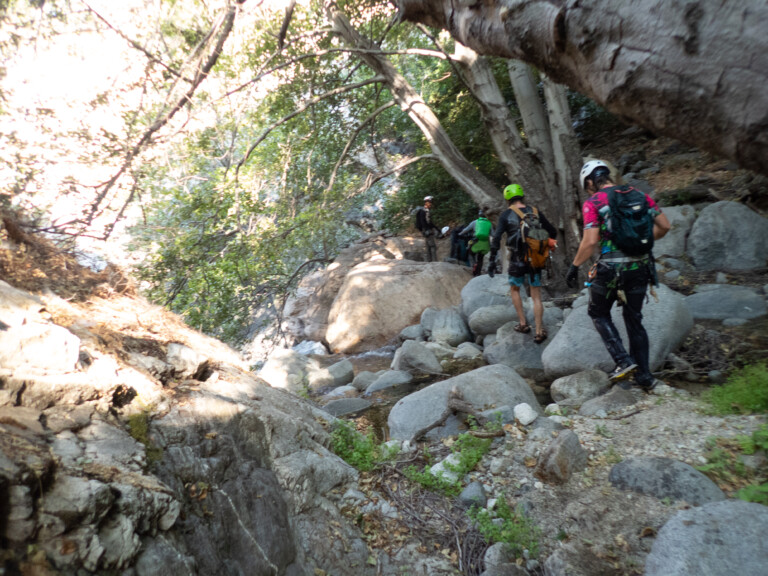 Several people equipped with helmets, backpacks, and outdoor gear walk along a rocky path and shallow stream surrounded by large boulders and tall trees in a forested canyon area, participating in a canyoneering activity.