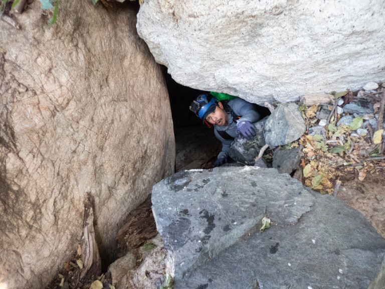 A canyoneer wearing a blue helmet with a mounted camera and gloves is climbing out of a narrow crevice between large rocks surrounded by dry leaves and stones in Eaton Canyon.