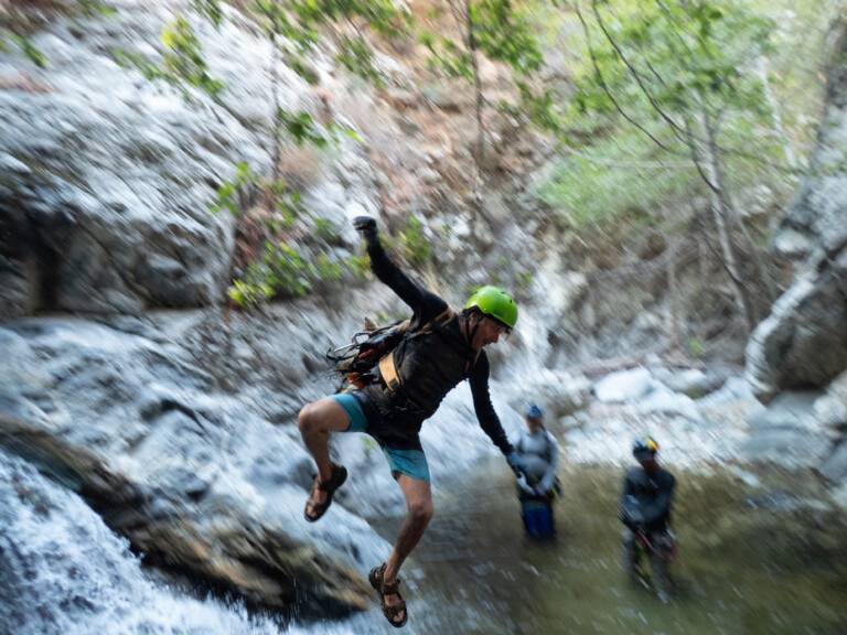 A person in a green helmet, black long-sleeve shirt, and blue shorts is mid-air jumping into a river surrounded by rocky canyon walls and greenery. Two other people wearing helmets and outdoor gear stand in the water in the background. The scene captures an adventurous moment during a canyoneering trip at Eaton Canyon.