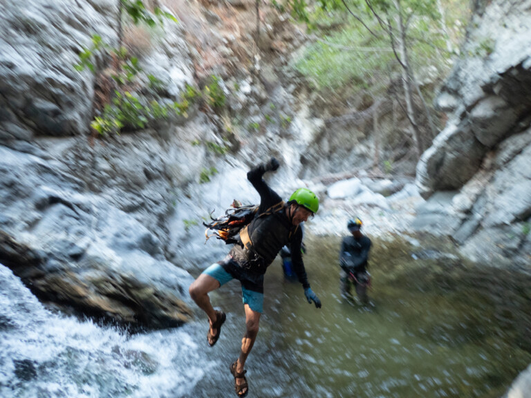 A person in a green helmet and canyoneering gear is mid-air jumping into a pool of water in a rocky canyon with trees and cliffs around. Another person stands in the water in the background, also wearing canyoneering gear.