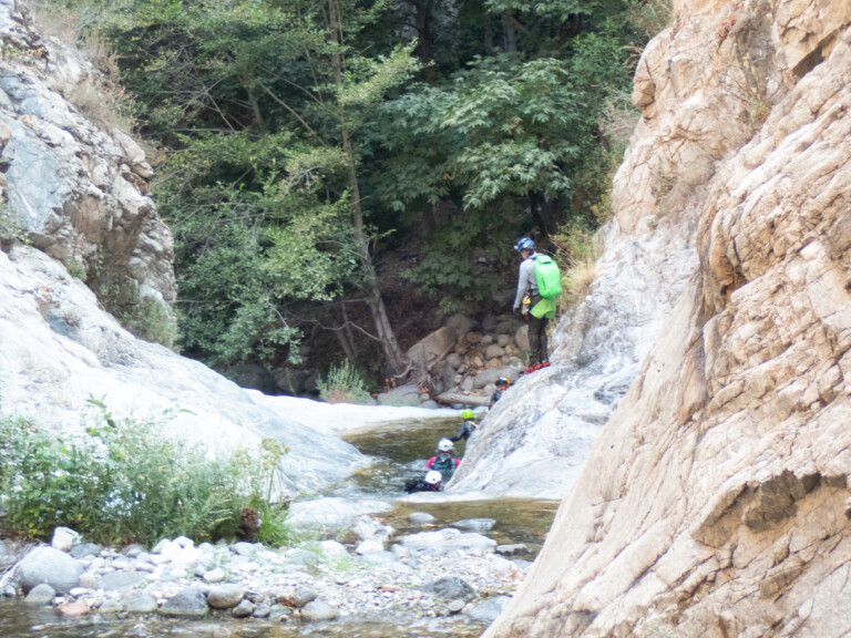Several people wearing helmets and outdoor gear are canyoneering in Eaton Canyon. They are moving through a shallow stream bordered by large rocky cliffs and dense green trees. One person with a bright green backpack stands on a rock overlooking the group below.