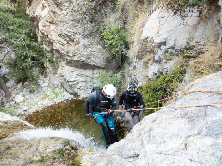 Two people equipped with helmets, harnesses, and ropes are rappelling down a rocky cliff next to a small waterfall and a shallow pool of water surrounded by rugged canyon walls and vegetation.