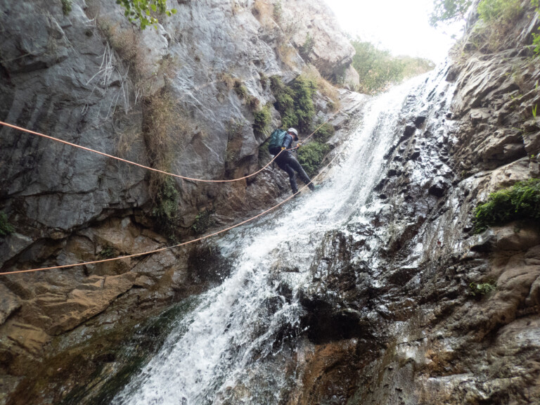 A person equipped with a helmet, backpack, and outdoor gear is rappelling down a steep waterfall in a rocky canyon. The individual is secured by orange ropes and surrounded by rugged rock formations and flowing water, with some greenery visible at the top edges of the canyon.