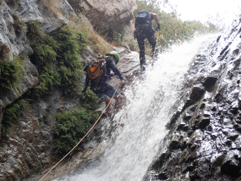 Two individuals engaged in canyoneering, descending a steep rocky waterfall in Eaton Canyon. They are wearing helmets, backpacks, and harnesses, using ropes for safety as water splashes around them. The surrounding area features rugged rocks and green vegetation.