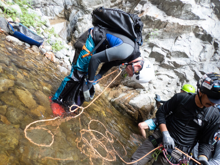 Two people wearing helmets and wetsuits are canyoneering in Eaton Canyon. One person is bent over near a rocky wall, holding onto a rope, while the other is sitting on a rock, also holding a rope. They are surrounded by rocky cliffs and shallow water with visible stones beneath the surface.