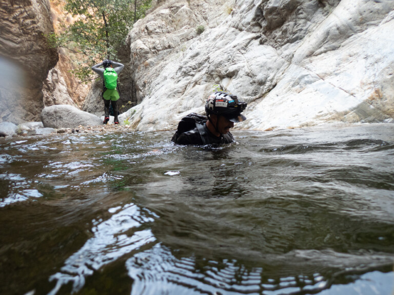 Two individuals canyoneering in Eaton Canyon; one person is partially submerged in water wearing a helmet with stickers and a black outfit, while the other stands on rocky terrain with a bright green backpack and outdoor gear, surrounded by rocky canyon walls and some greenery.