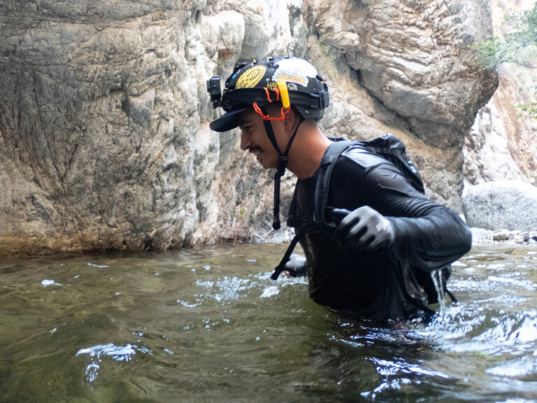 A man dressed in black with a helmet and gloves is wading through shallow water surrounded by rocky canyon walls, engaged in canyoneering at Eaton Canyon.