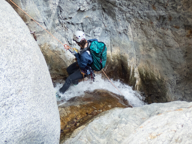 A person equipped with a white helmet, green backpack, and climbing gear is rappelling down a rocky waterfall in Eaton Canyon. The individual is holding onto an orange rope secured to the rock face, surrounded by rugged gray rocks and flowing water below.