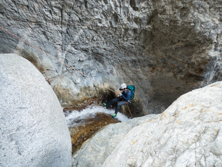 A person equipped with a white helmet, teal backpack, and outdoor gear is rappelling down a steep rocky canyon wall using an orange rope. They are descending into a shallow stream of water flowing over rocks in Eaton Canyon, surrounded by rugged gray rock formations.