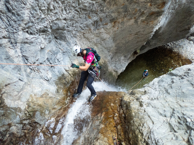 Two individuals wearing helmets and climbing gear rappelling down a steep rocky waterfall in Eaton Canyon. One person is closer to the camera descending the wet rock face, while the other is further below near a pool of water, both connected by orange ropes.