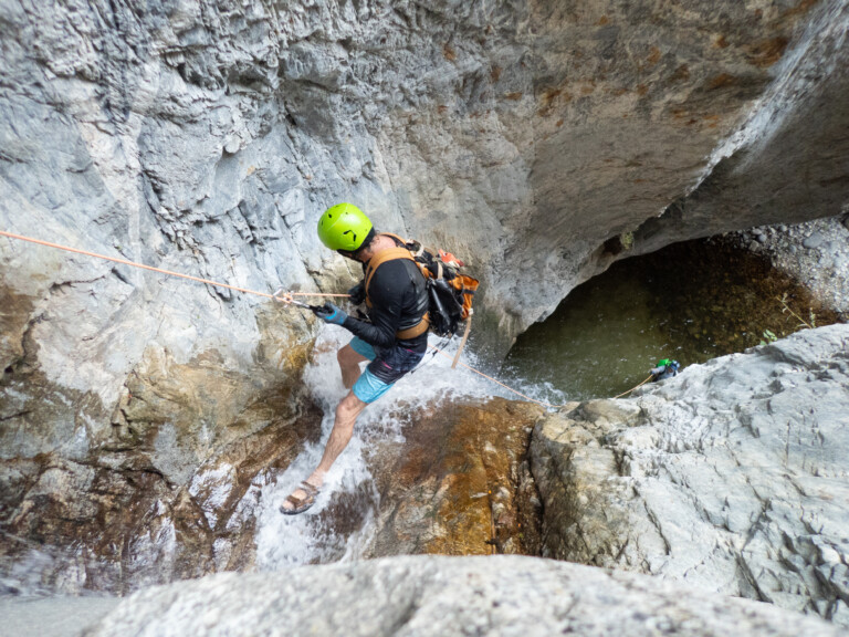 A person with a bright green helmet, black long-sleeve shirt, blue shorts, and gloves rappelling down a rocky waterfall using an orange rope in a narrow canyon. The rocky walls surround a small pool of water below, and another person is visible further down the canyon also engaged in canyoneering.