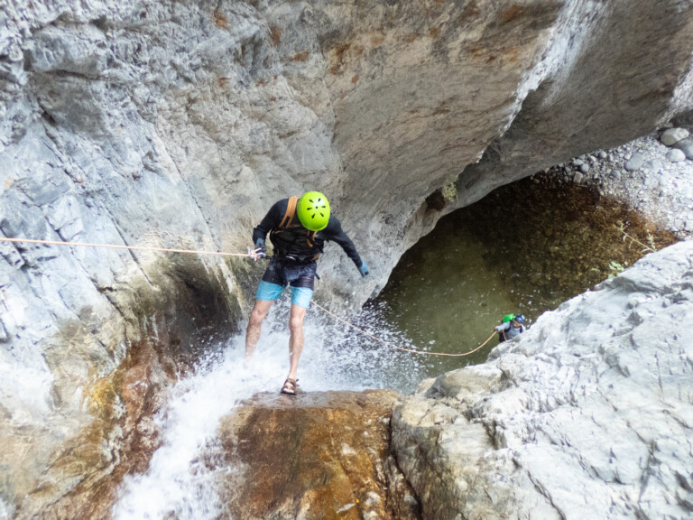 Two individuals engaged in canyoneering, rappelling down a steep rocky waterfall in Eaton Canyon. The person in the foreground wears a bright green helmet, black long-sleeve shirt, and blue shorts, descending with a rope over wet rocks and splashing water. Another person is visible below near a pool of water surrounded by rocks.
