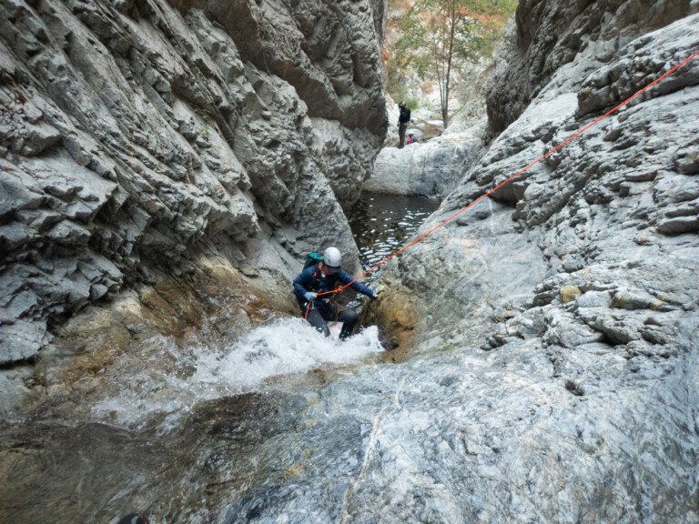 A person in outdoor gear and helmet rappels down a rocky waterfall in a narrow canyon with steep gray rock walls. An orange rope is secured to the rocks, and two other people stand on rocks near a pool of water in the background. Sparse trees with green and orange leaves are visible beyond the canyon walls.