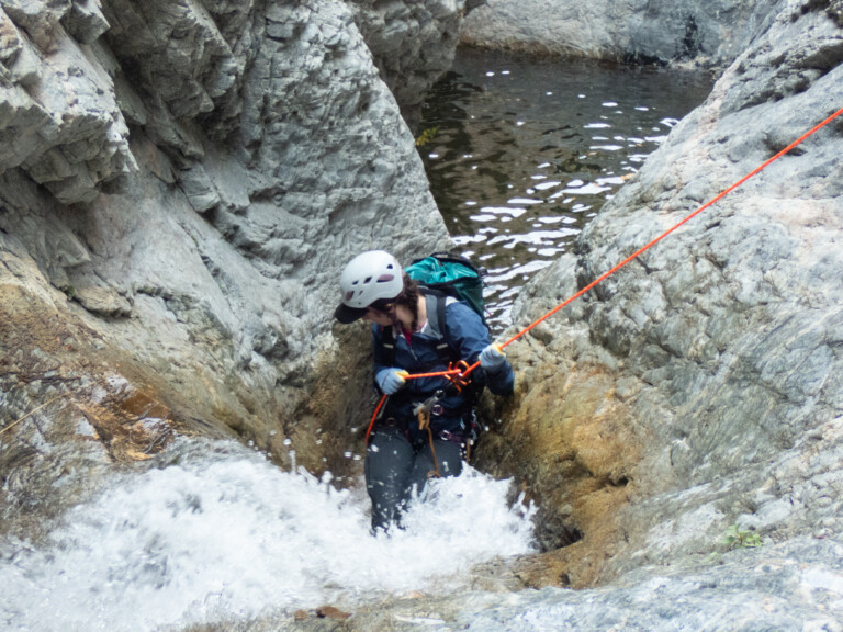 A person equipped with a white helmet, blue jacket, gloves, and a backpack is rappelling down a rocky waterfall in Eaton Canyon. They are holding an orange rope and descending carefully amidst flowing water and rugged rock formations.
