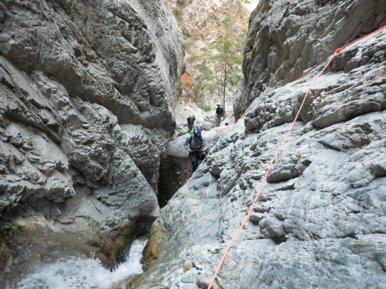 Several people wearing helmets and backpacks rappelling down a narrow rocky canyon with steep walls and a small pool of water below, using bright orange safety ropes. The canyon is surrounded by rugged rock formations and sparse vegetation.