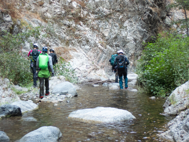 Five people equipped with helmets, backpacks, and outdoor gear walking through a shallow stream in a rocky canyon with green bushes and trees on the sides, under overcast sky conditions.
