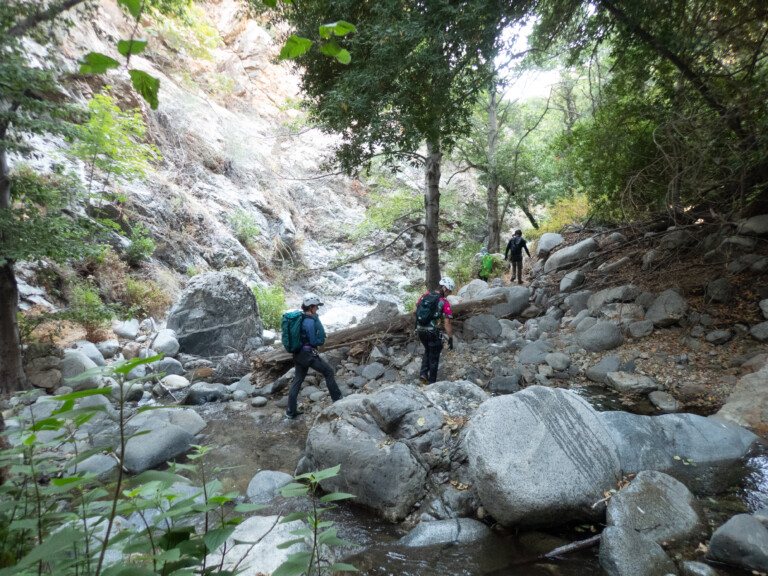 Several people equipped with helmets and backpacks are hiking through a rocky canyon with a shallow stream, surrounded by trees and steep rock walls in Eaton Canyon.