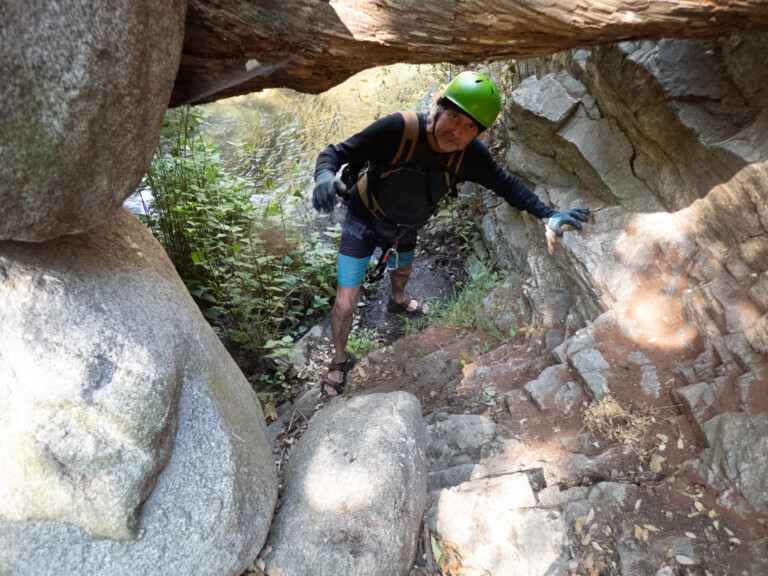 A man with a green helmet, black long-sleeve shirt, blue shorts, and gloves climbs through a narrow rocky canyon path beside a flowing stream surrounded by green plants and large boulders.