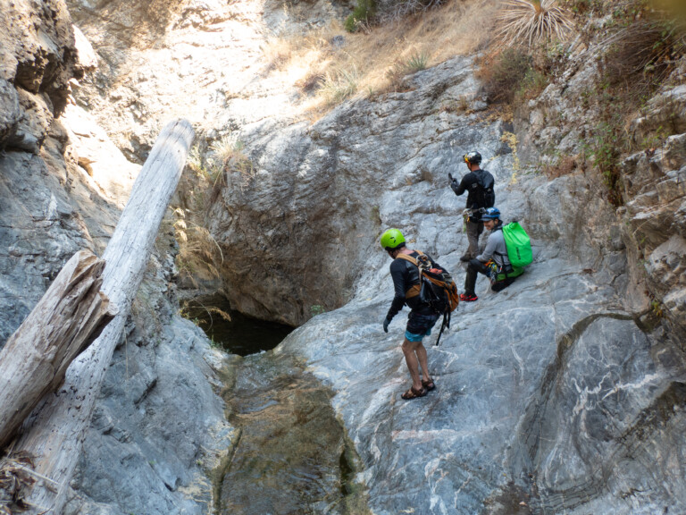 Three people wearing helmets and outdoor gear are canyoneering in a rocky canyon with dry vegetation. One person is standing near a small pool of water, another is sitting on a rock with a bright green backpack, and the third is preparing to descend the rocky slope. The scene is illuminated by natural sunlight highlighting the rugged rock formations and dry plants.