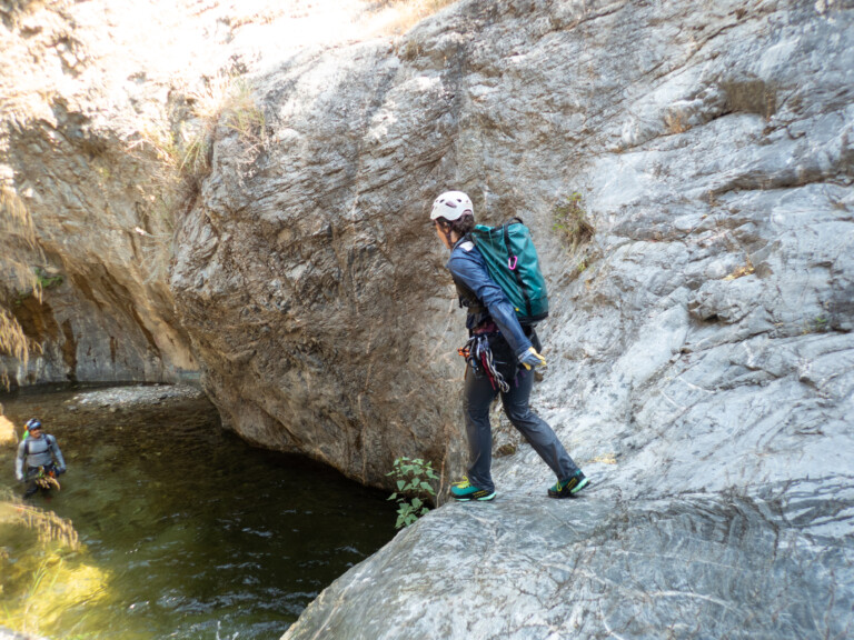 A person wearing a white helmet, blue jacket, black pants, and green shoes stands on rocky terrain near a water pool in Eaton Canyon. Another person wearing a helmet and outdoor gear is seen in the water below, surrounded by steep rock walls under bright sunlight.