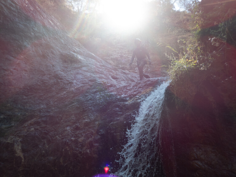 A person equipped with a helmet and harness is canyoneering down a rocky waterfall in Eaton Canyon. The scene is illuminated by intense sunlight from above, causing glare and lens flare. Surrounding the waterfall are rocks and vegetation.