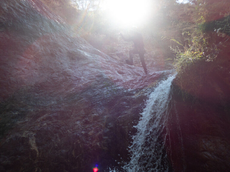 A person wearing canyoneering gear climbs up a rocky waterfall in Eaton Canyon. Bright sunlight shines from above, creating a strong lens flare and illuminating water droplets around the climber. Surrounding the waterfall are rocks and some vegetation.