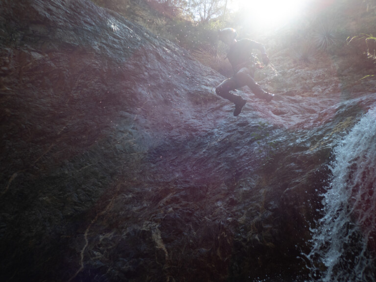 A person equipped with a helmet and harness is rappelling down a wet rocky cliff face in Eaton Canyon. Sunlight shines brightly from the top right corner, illuminating water droplets falling alongside the person. The surrounding area features rugged rock formations and sparse vegetation.