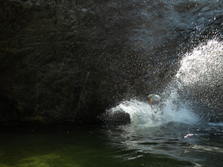 A person wearing a helmet is splashing into water inside a dark rocky canyon, with water droplets scattering in the air around them.