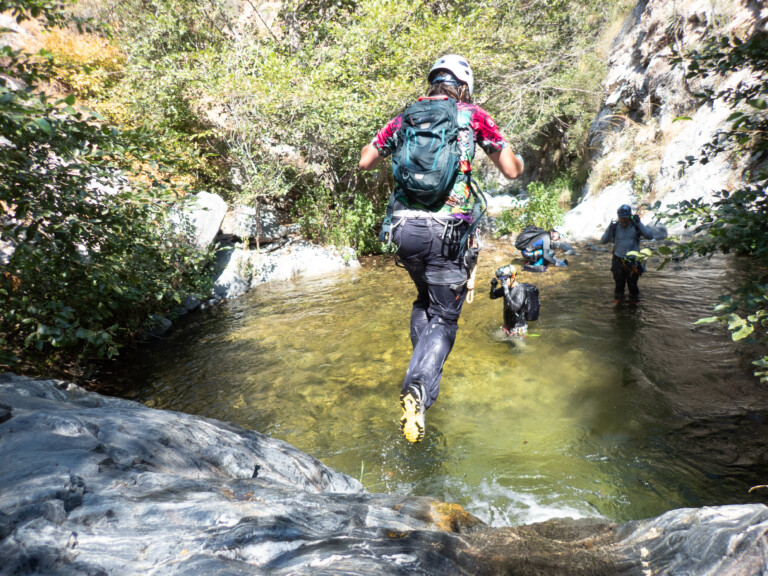 A person wearing a white helmet, colorful shirt, and backpack is stepping into a shallow stream surrounded by rocks and greenery. Two other people in helmets and outdoor gear are also in the water, preparing for canyoneering in a rocky canyon environment with sunlight filtering through the trees.