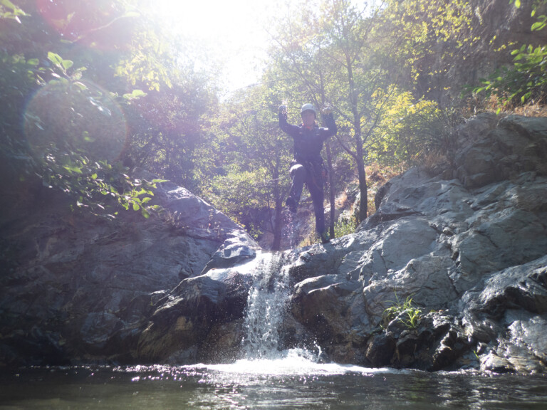 A person in canyoneering gear and helmet is descending a rocky waterfall surrounded by lush green trees and bright sunlight in Eaton Canyon. Water splashes down the rocks into a pool below, with sunlight creating lens flare effects.