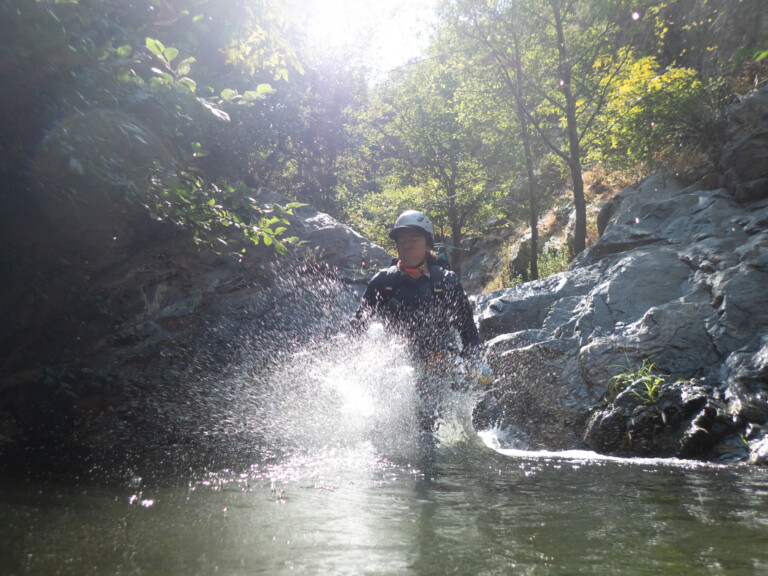A person in a helmet and outdoor gear is splashing water while canyoneering in a rocky canyon area with green trees and bright sunlight in the background.