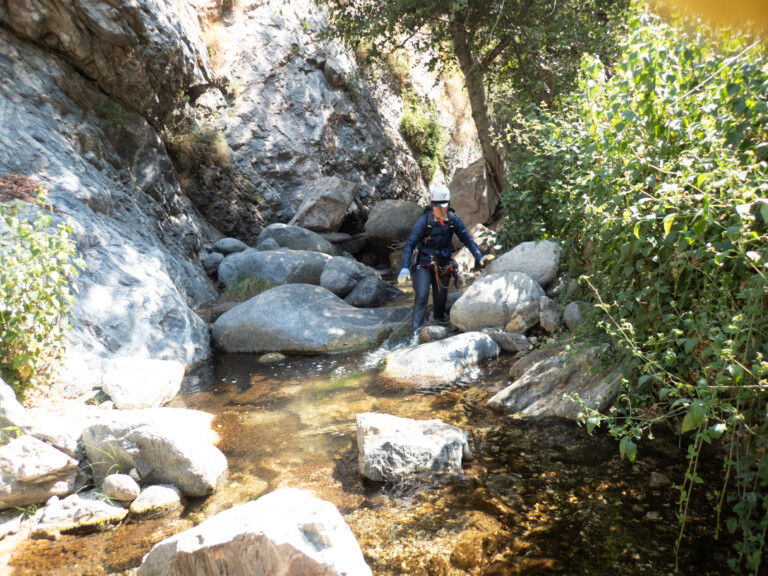 A person dressed in canyoneering gear including a helmet, gloves, and harness walks carefully through a shallow rocky stream surrounded by large boulders and green foliage in Eaton Canyon under bright daylight.