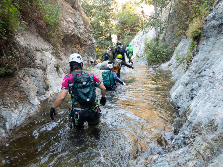 Several people equipped with helmets, backpacks, and gloves wading through a shallow stream surrounded by rocky canyon walls and greenery in Eaton Canyon, engaging in canyoneering.