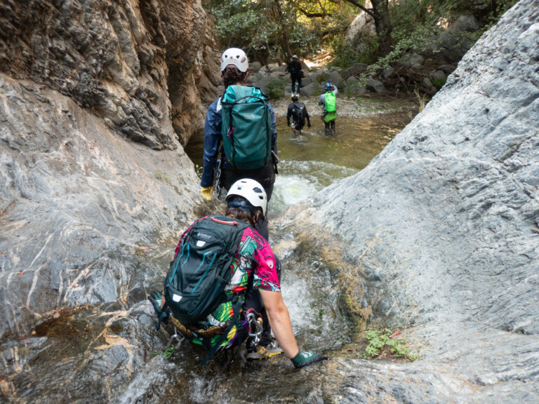 Several people wearing helmets and backpacks are canyoneering in Eaton Canyon, walking through a shallow stream surrounded by rocky cliffs and green trees, with one person in the foreground carefully descending a rock slope into the water.