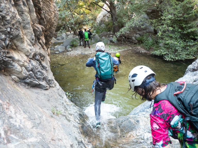 Several people equipped with helmets, backpacks, and outdoor gear are canyoneering in Eaton Canyon. They are crossing a shallow stream surrounded by large rocks and dense green foliage, with one person stepping into the water and others ahead on the rocky path.