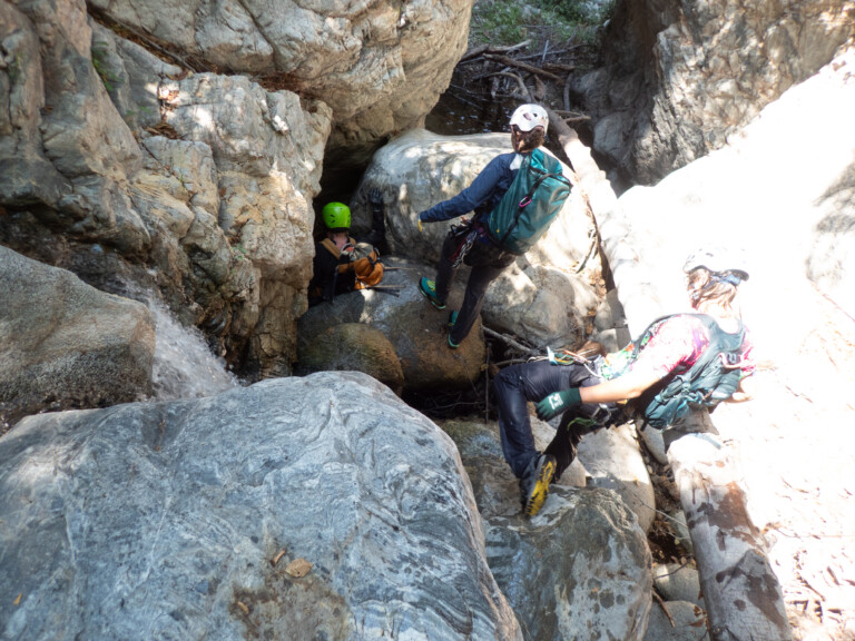 Three individuals equipped with helmets, harnesses, and backpacks are engaged in canyoneering in a rocky canyon. One person with a green helmet is descending into a narrow crevice near water, while another with a white helmet and teal backpack stands on a rock, and a third person with a white helmet and colorful sleeves is preparing to descend. The scene is surrounded by large rocks and some scattered branches.
