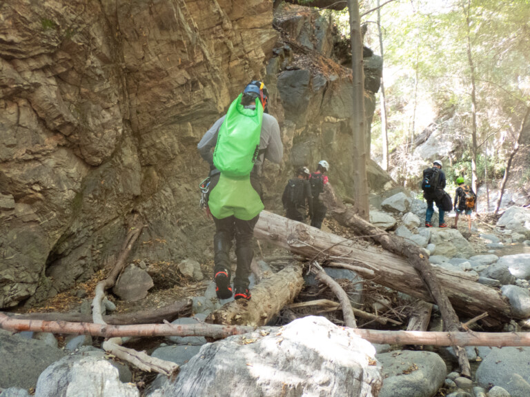 Several people wearing helmets and carrying backpacks walk along a rocky trail in a canyon surrounded by large rock walls and trees, with sunlight filtering through the foliage.