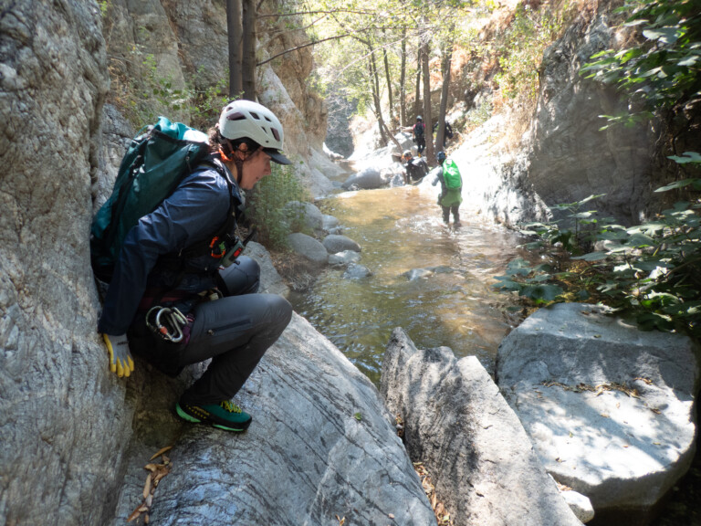 A person wearing a white helmet, blue jacket, and green shoes crouches on a rock beside a narrow stream in a rocky canyon. In the background, three other people with helmets and backpacks are walking through the shallow water surrounded by tall rock walls and trees with sunlight filtering through the leaves.