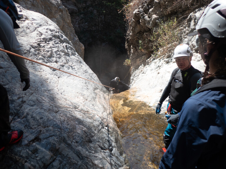 Several people equipped with helmets, wetsuits, and gloves are engaged in canyoneering, rappelling down a steep rocky canyon with a small stream of water flowing through it. The scene is set in Eaton Canyon with rugged rock formations and some vegetation visible on the canyon walls.