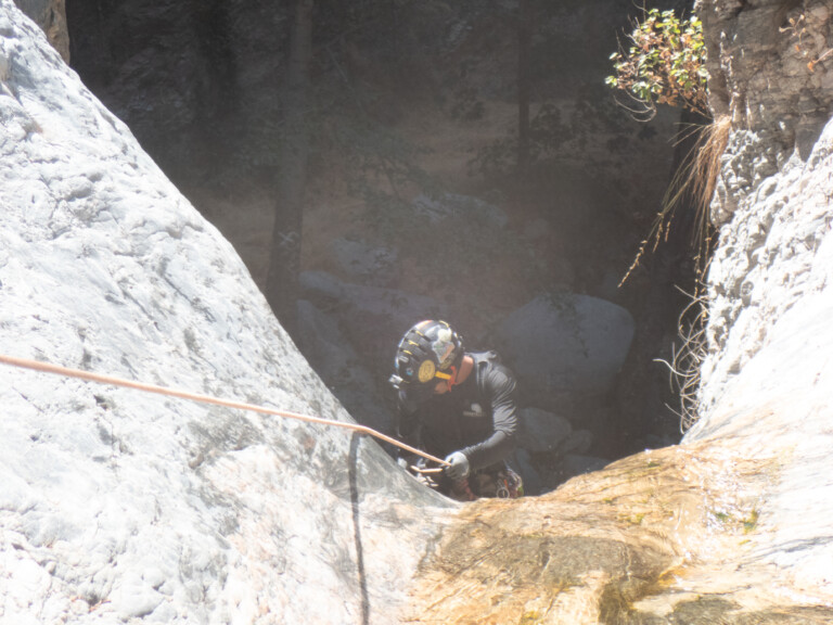 A person equipped with a helmet and gloves is descending a steep rocky canyon wall using a rope, surrounded by large rocks and sparse vegetation in Eaton Canyon.