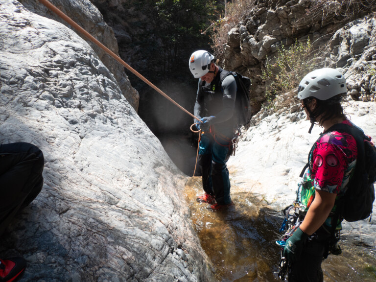 Two individuals equipped with helmets, harnesses, and gloves are engaged in canyoneering in Eaton Canyon. One person is standing in shallow water holding a rope, preparing for descent, while the other watches attentively. They are surrounded by steep rocky canyon walls and sparse vegetation under bright daylight.