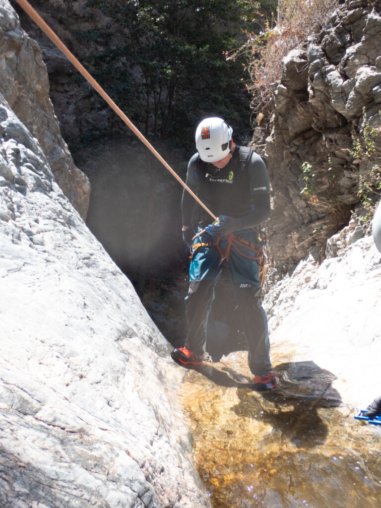 A person dressed in canyoneering gear including a white helmet with an Uber Adventures sticker, gloves, and harness, is rappelling down a rocky canyon wall using a rope. The scene is set in Eaton Canyon with rocky cliffs and some vegetation in the background, and water flowing at the person's feet.