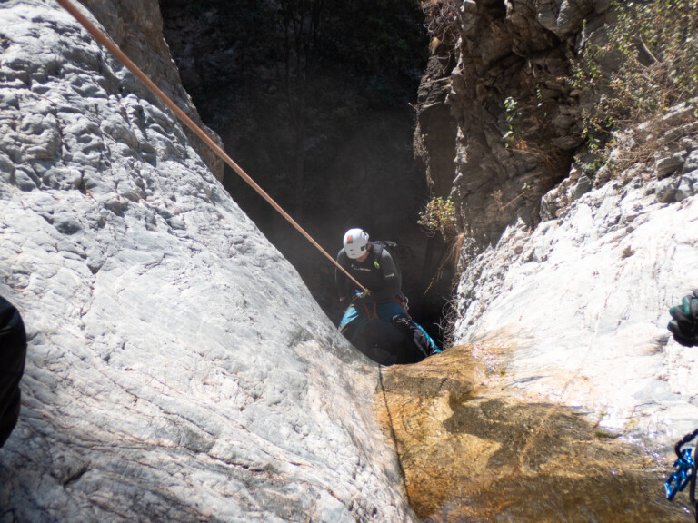 A person equipped with a white helmet, black long-sleeve shirt, and blue pants is rappelling down a steep rocky canyon wall using a rope. The canyon walls are rugged with some vegetation, and there is a small stream of water flowing down the rocks. The scene is illuminated by sunlight, highlighting the texture of the rocks and the natural environment of Eaton Canyon.