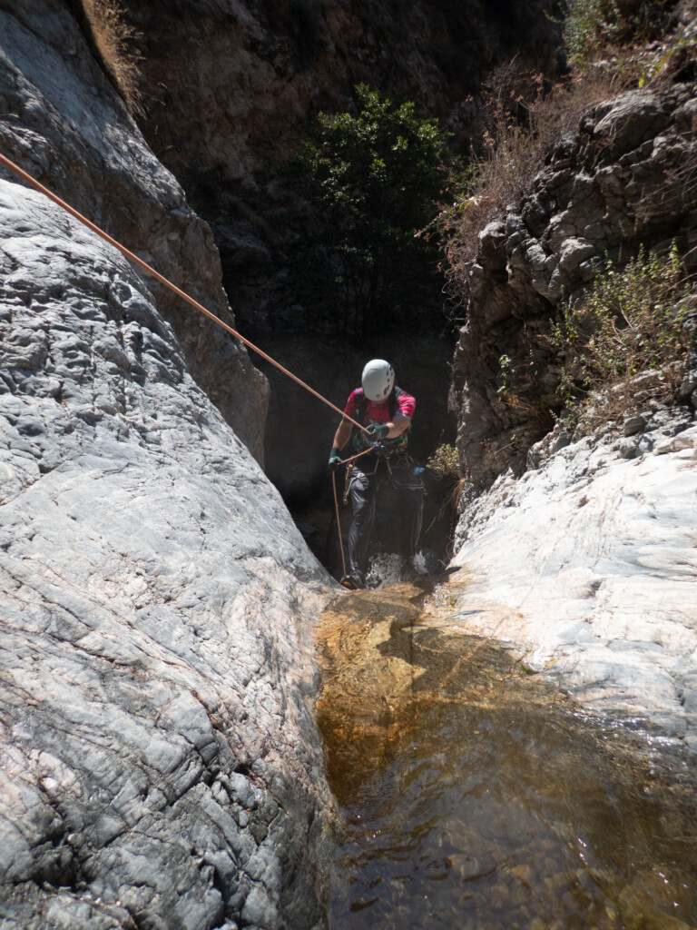 A person equipped with a white helmet, gloves, and harness is rappelling down a steep rocky canyon wall using a rope. The canyon features rugged gray rocks and a small stream of water flowing at the bottom. Sparse bushes and plants grow on the rocky terrain under bright daylight.